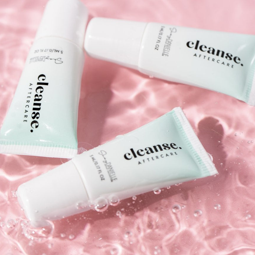 Three Girlz Ink Cleanse Aftercare tubes displayed on a pink water background, designed for gentle cleansing and healing after permanent makeup or tattoo procedures.