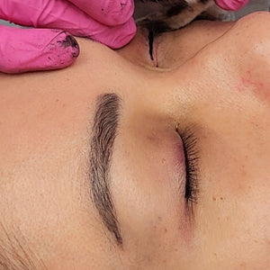 Eyebrow tattoo touch-up performed with OMG® Pigments Bitchin Brown by Teryn Darling. The image shows healed, natural-looking brows one year post-procedure, with the artist using pink gloves during the session.