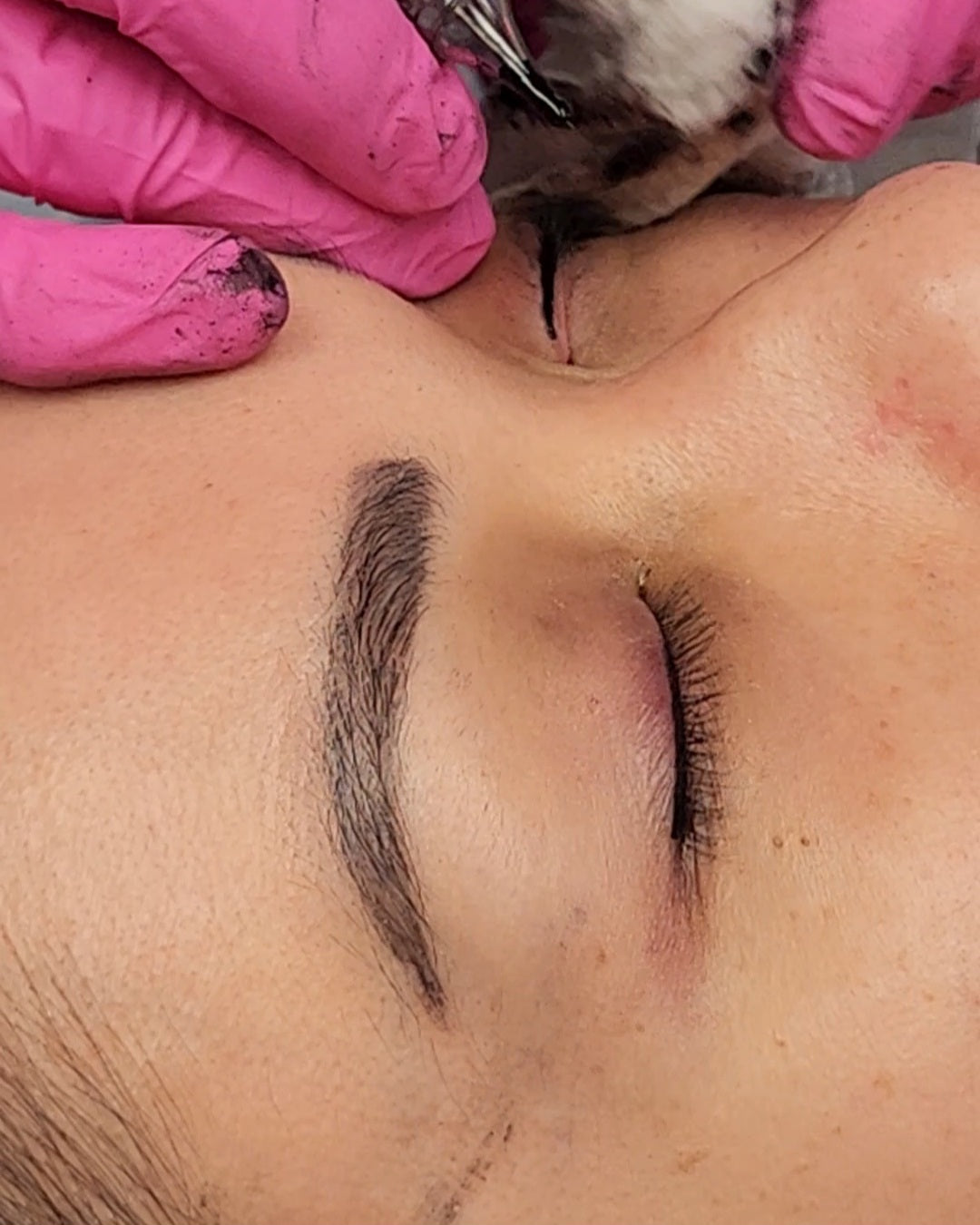 Eyebrow tattoo touch-up performed with OMG® Pigments Bitchin Brown by Teryn Darling. The image shows healed, natural-looking brows one year post-procedure, with the artist using pink gloves during the session.