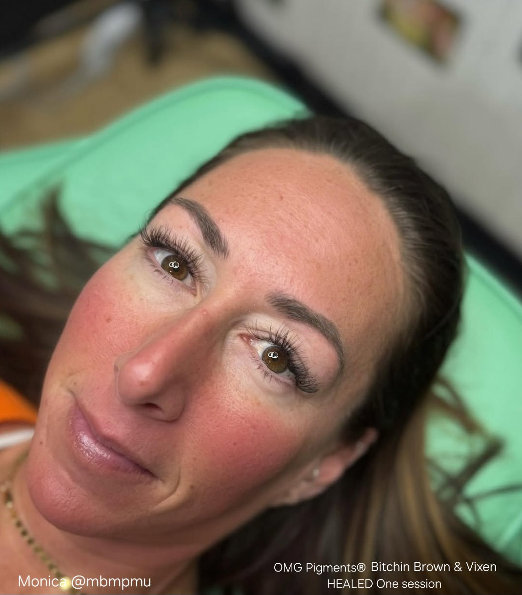 Healed brow results after one session using OMG® Pigments in Bitchin Brown and Vixen. The image shows a woman lying on a green surface with soft, natural brow color retention created by Monica @mbmpmu.
